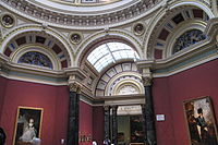 Octagonal room with rich red wallpaper, paintings and architectural details in marble of various colours