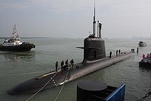 A Scorp&egrave;ne class submarine at dock, half out of the water. People on top are mooring it, and a boat can be seen in the background